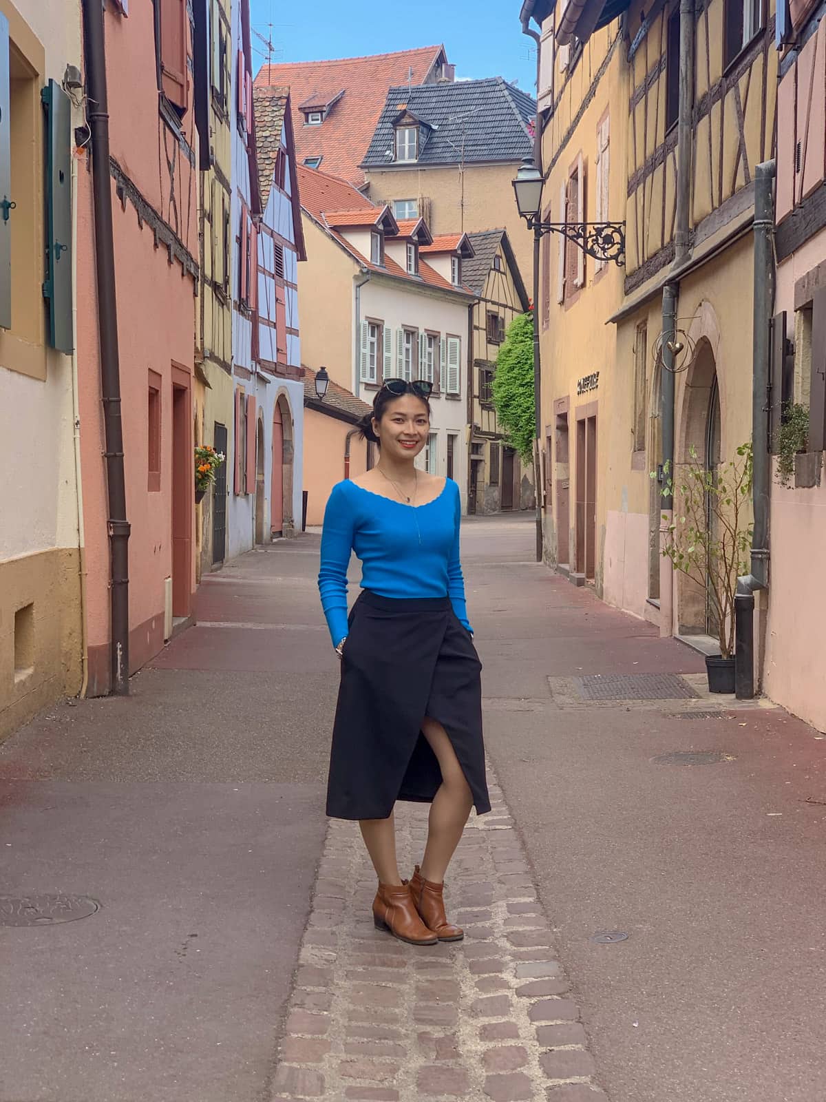 A woman in a bright blue top and a black skirt, with her hands in her pockets. She is wearing brown shoes and standing in a small alleyway between some old pink buildings