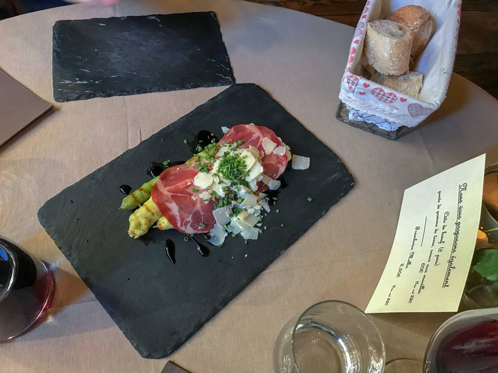 Asparagus with a slice of proscuitto and shaved cheese, presented on a black stone board on a dinner table in a restaurant