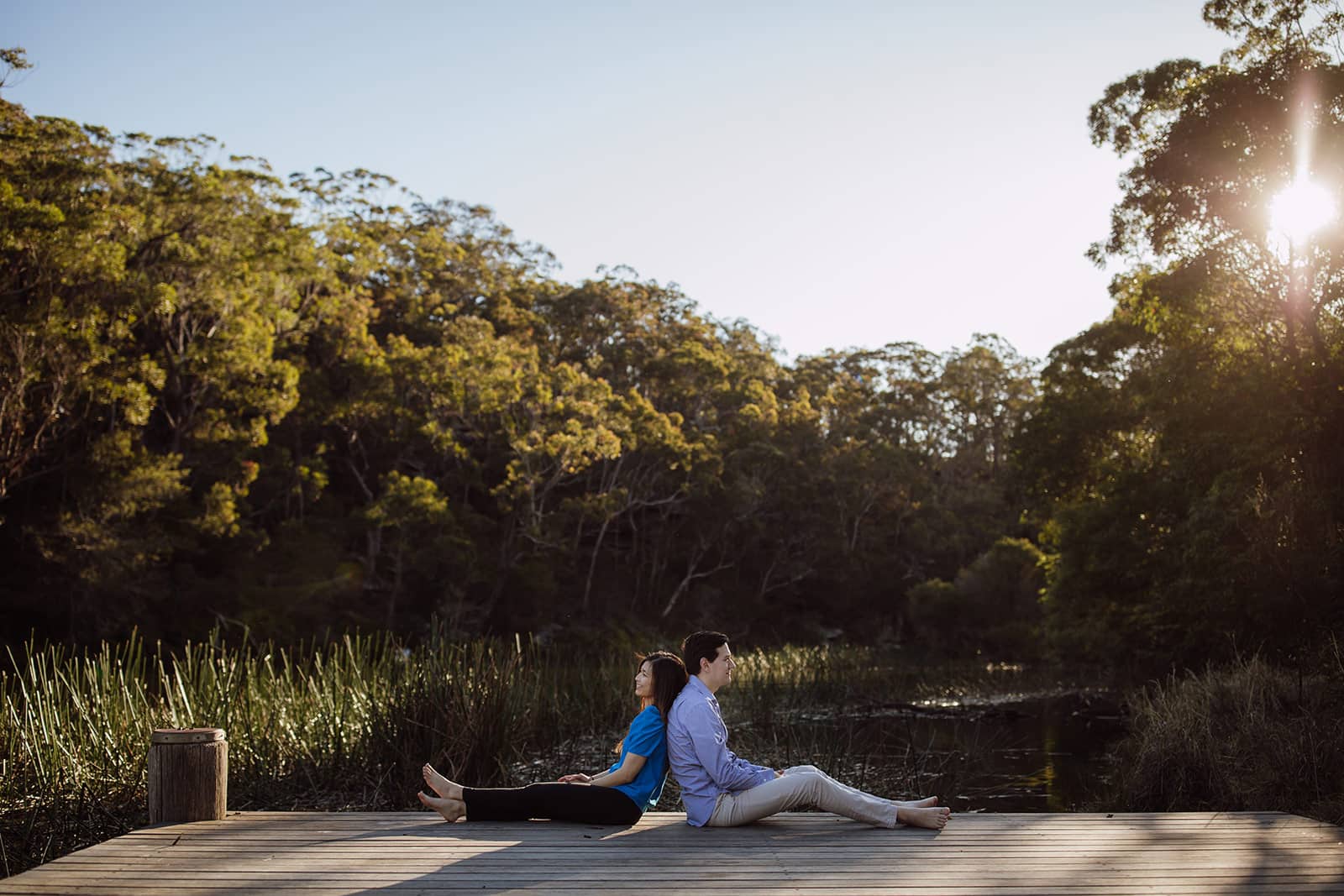 A man and woman sitting back-to-back on a pier, facing away from each other and smiling.