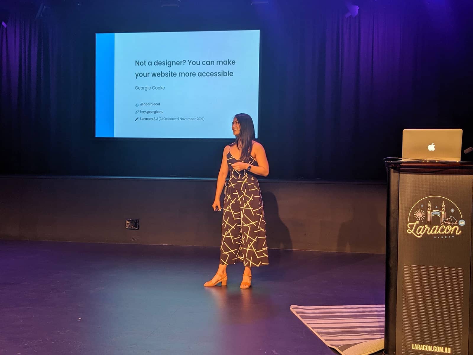 A woman in a black patterned jumpsuit presenting with a stage behind her that has presentation slides on it.