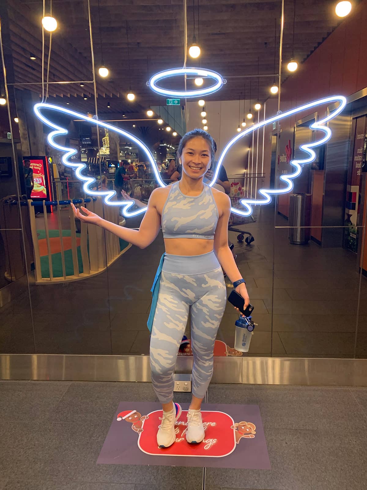A woman in a sports bra and leggings, holding a bottle in one hand and holding one hand up in the air. She is in front of a backdrop with neon lighting arranged like angel wings