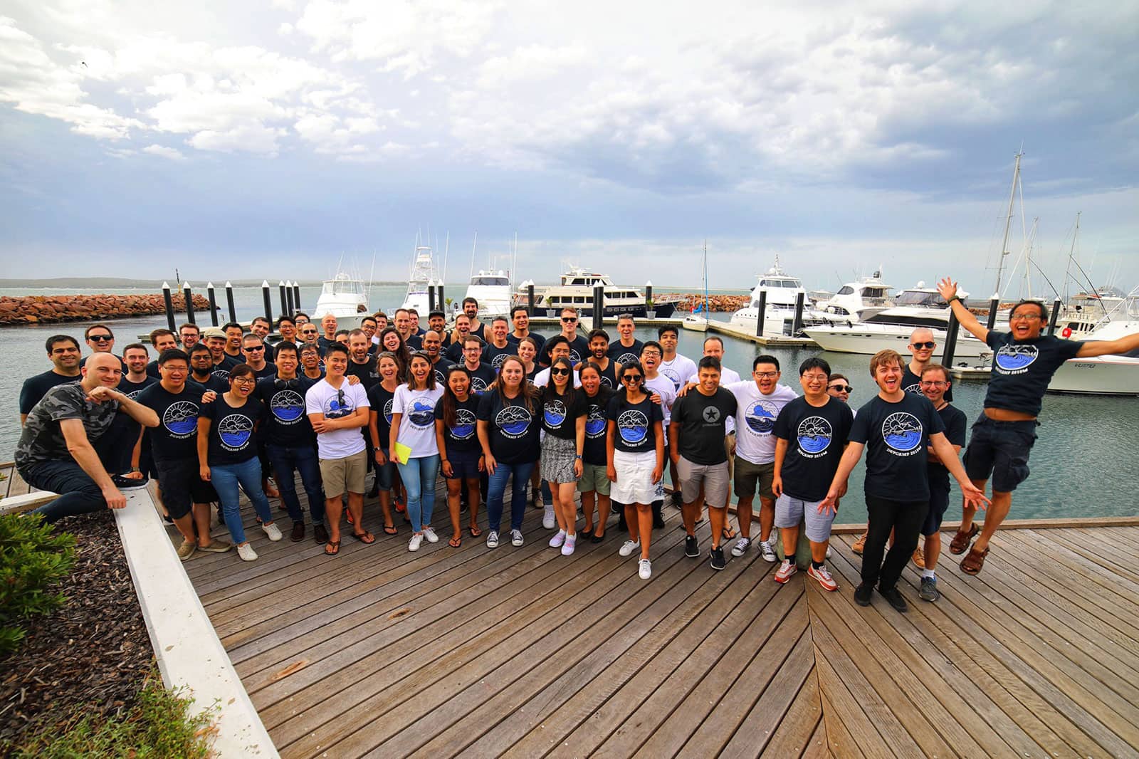 A group of people, all smiling or looking excited, standing on a boardwalk with some boats in the background. The people are wearing shirts with similar designs, mostly dark coloured but some white in colour