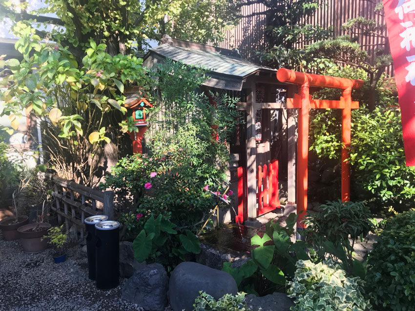 A Japanese shrine with an orange torii gate, a small water feature, a small wooden hut with an awning, and a lot of green leafy trees