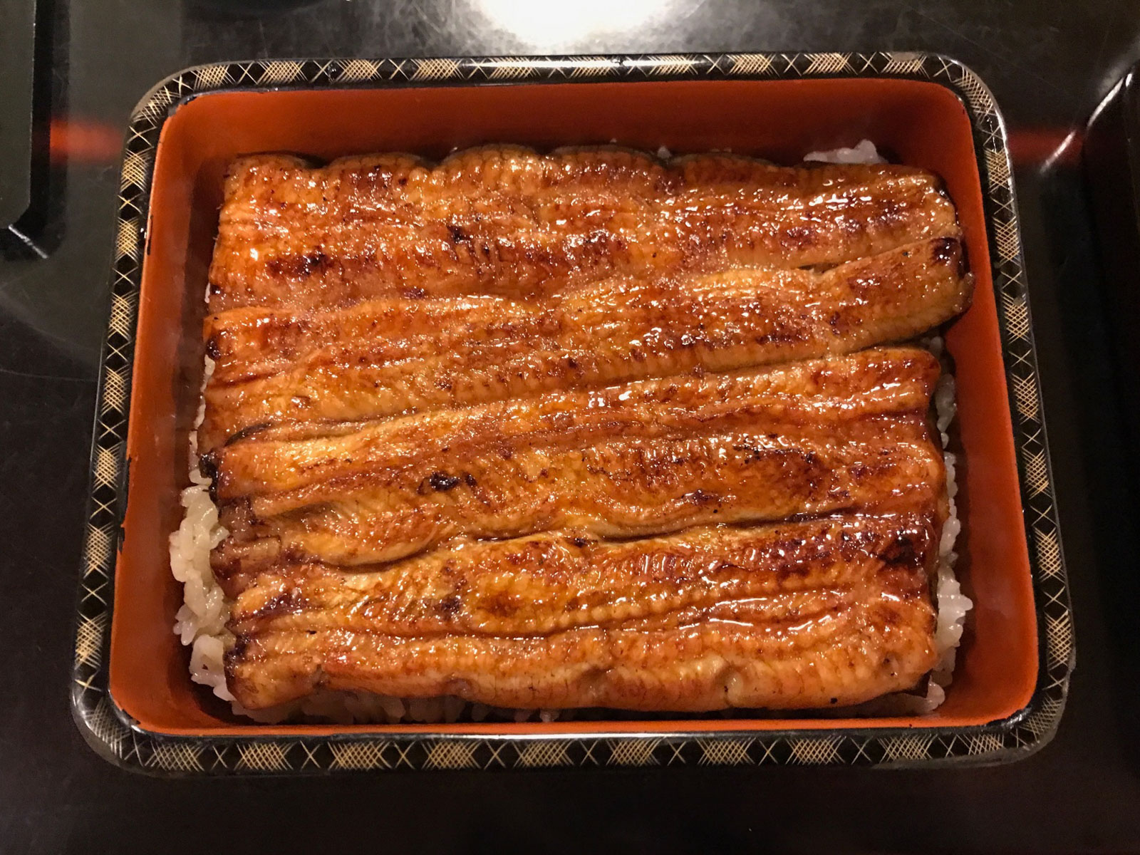 A rectangular box filled with slices of grilled eel, golden brown in colour. Some white rice can be seen underneath the eel. The interior of the box is a dark orange-red, and the rim has a gold and black pattern.