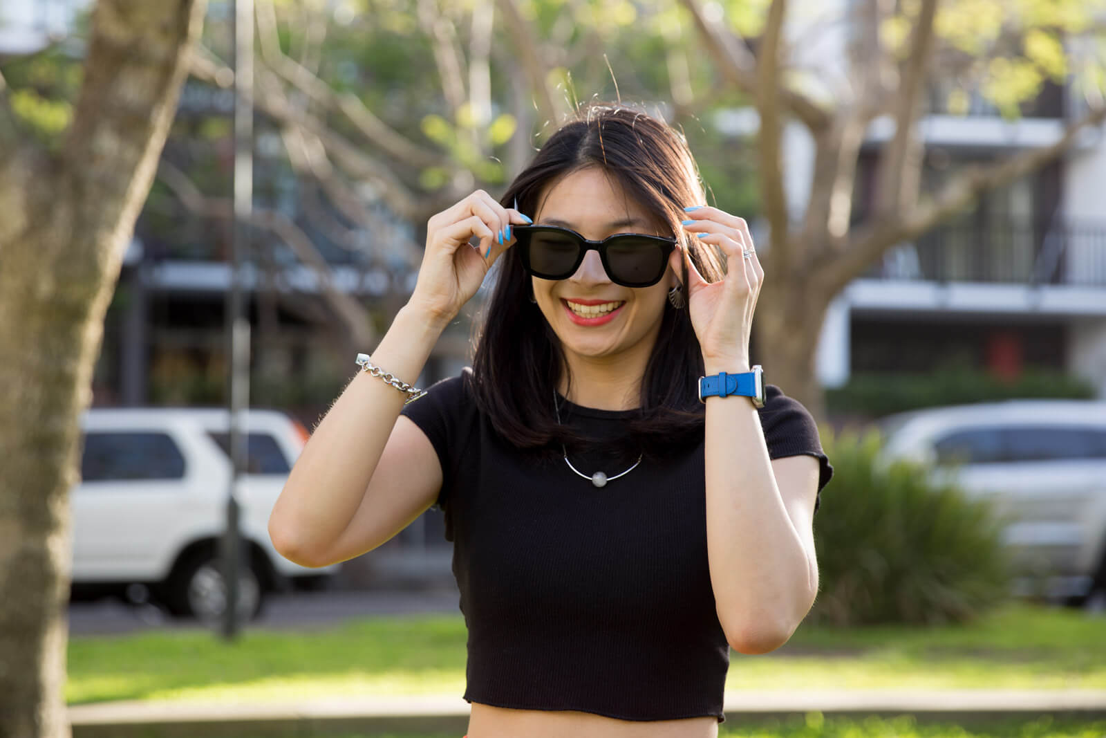 A medium shot of the same woman in previous photos on this page, midway through placing her black sunglasses on her face. She is grinning.