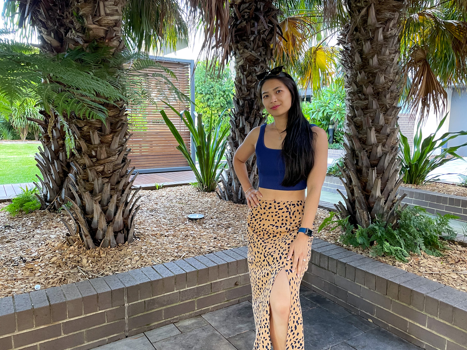 An Asian woman with long dark hair, wearing a long tan coloured midi skirt with a dark animal print, and a fitted blue top. She has a hand on her hip and sunglasses on top of her head. She is standing on concrete tiles and in the background are some planted trees.