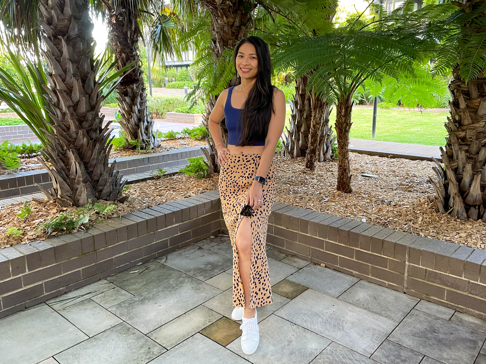 An Asian woman with long dark hair, wearing a long tan coloured midi skirt with a dark animal print, and a fitted blue top. She has a hand on her hip. She is standing on concrete tiles and in the background are some planted trees.