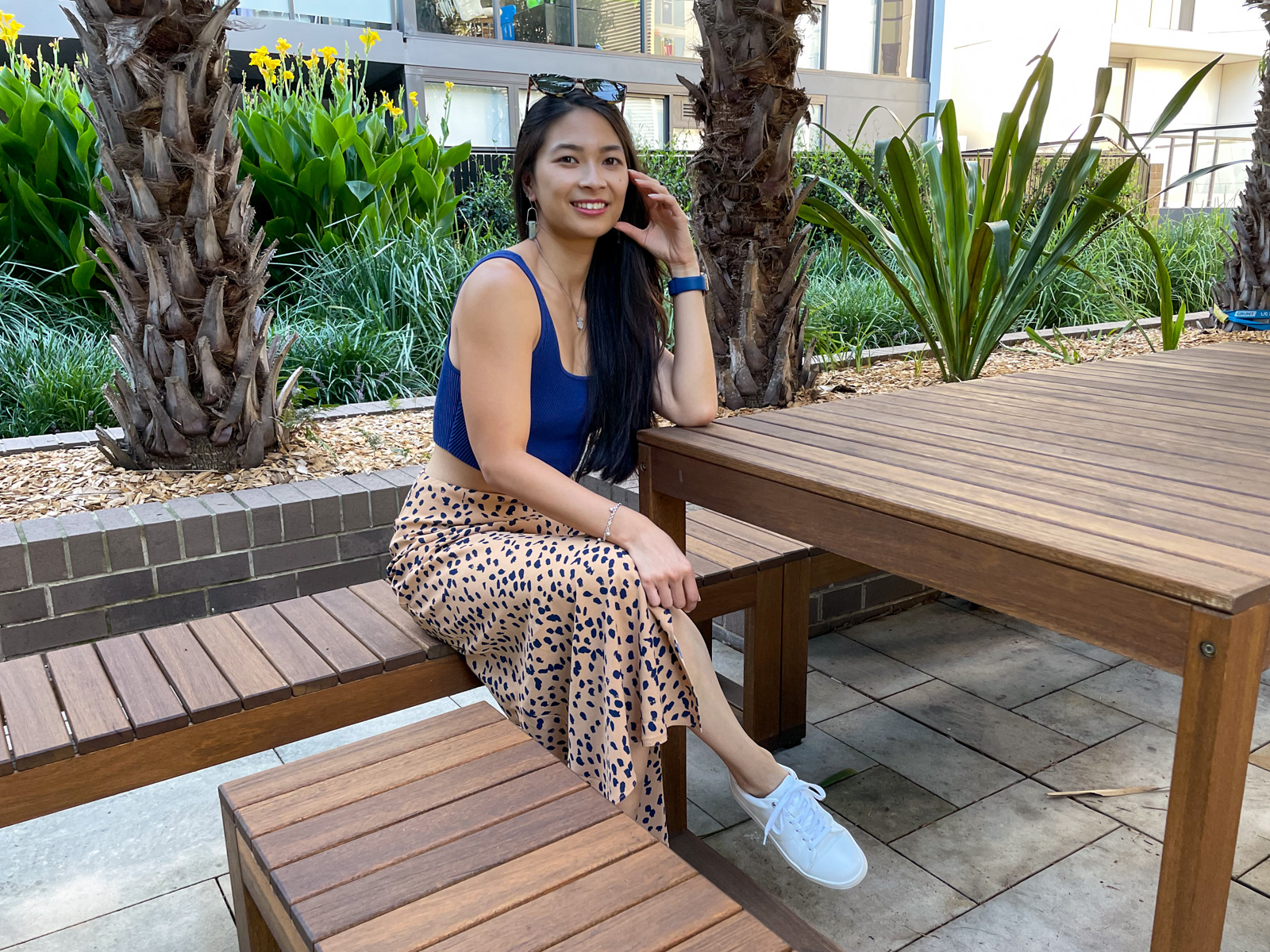 An Asian woman with long dark hair, wearing a long tan coloured midi skirt with a dark animal print, and a fitted blue top. She is sitting on a wooden bench alongside a wooden table and is leaning on the table with her elbow.