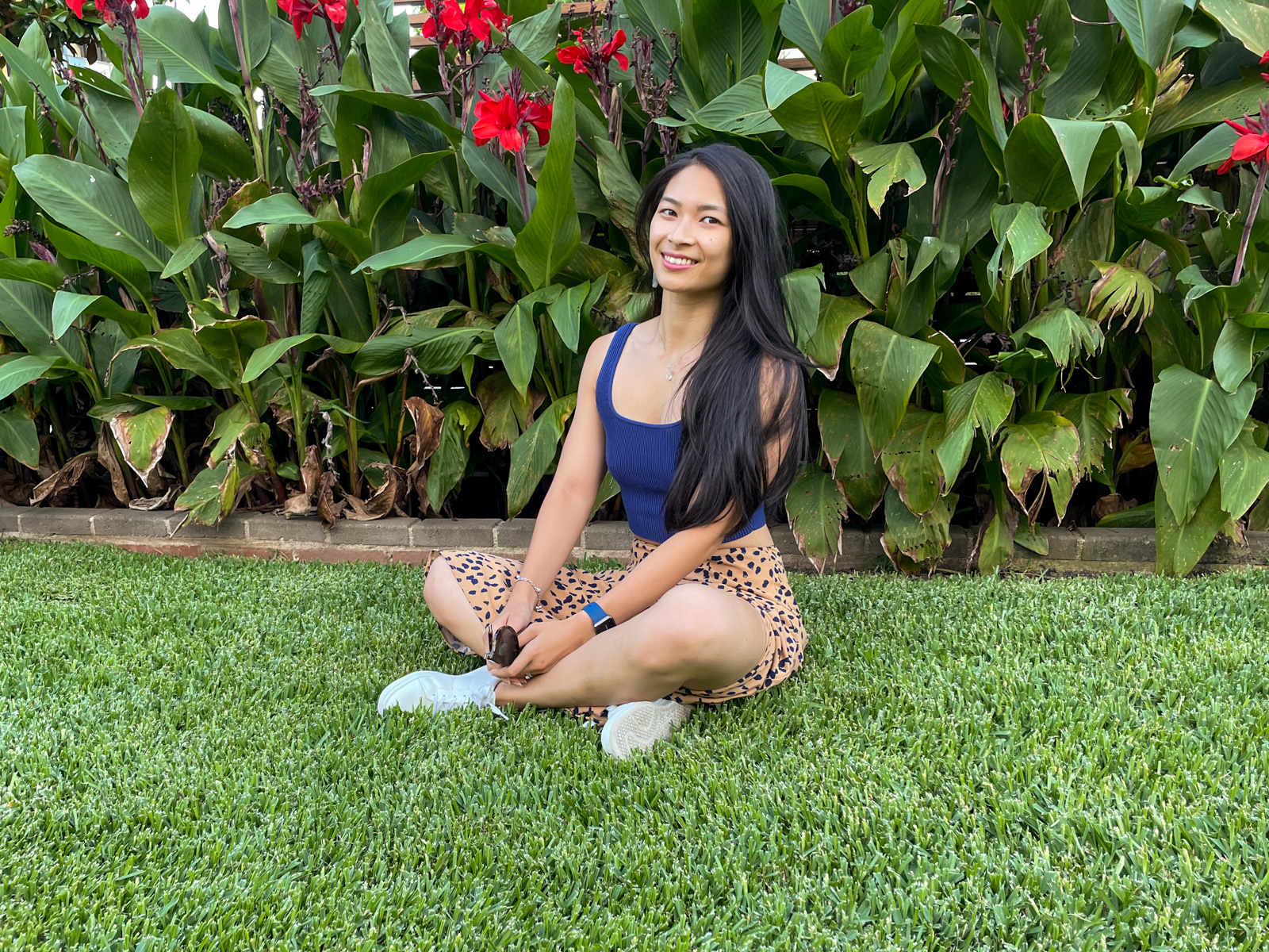 An Asian woman with long dark hair, wearing a long tan coloured midi skirt with a dark animal print, and a fitted blue top. She is sitting on freshly mowed green grass with her hands in her lap.