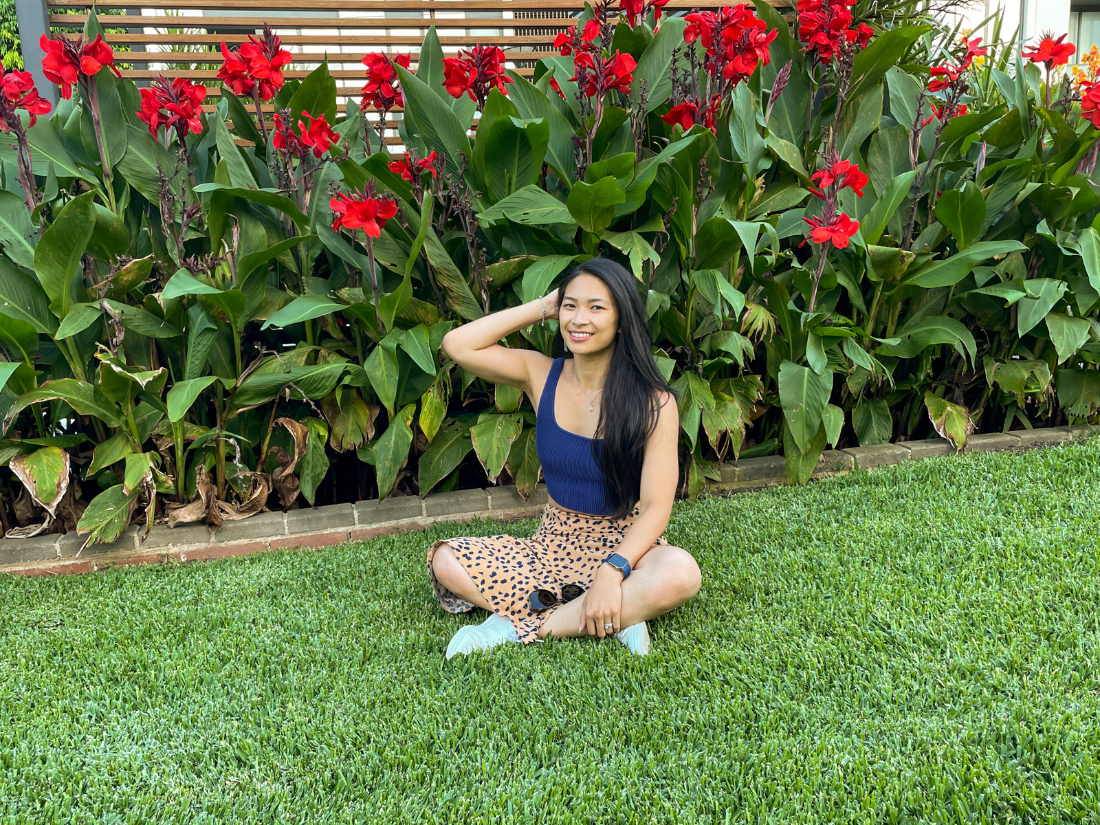 An Asian woman with long dark hair, wearing a long tan coloured midi skirt with a dark animal print, and a fitted blue top. She is sitting on freshly mowed green grass with a hand touching her hair as her elbow is bent. There are red flowers in the green plants behind her.