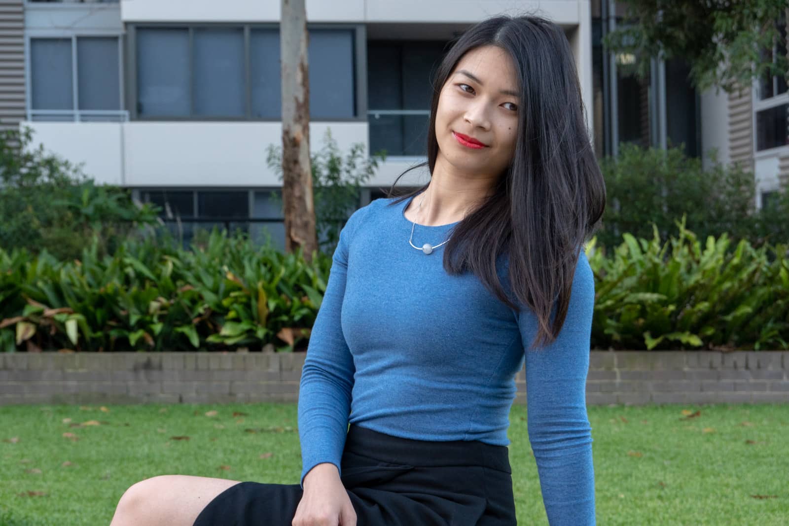 A medium shot of a woman with short dark hair, wearing a long-sleeved indigo blue top, looking slightly away from the camera. She has bright red-orange lipstick on and a curved necklace with a light grey bead in the middle. She is sitting in a grassy area with a residential building in the background.