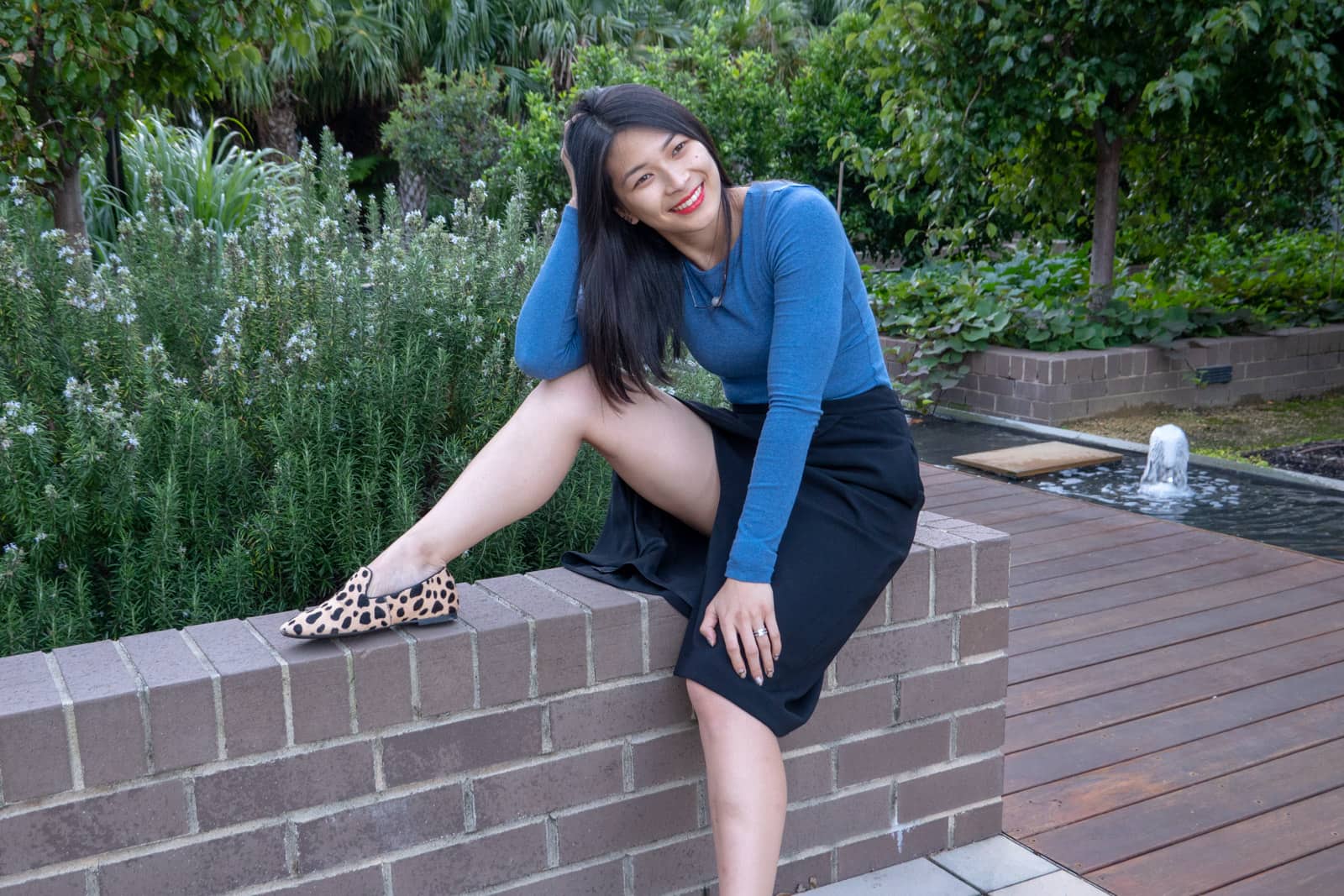 A woman with short dark hair, dressed in a blue top and a long black skirt, sitting on a short brick wall with one leg up on the wall and bent. Her head is resting on her palm with her elbow propped on one knee. Her other hand is on her other knee. She is wearing light tan loafers on her feet, that have black giraffe spots.