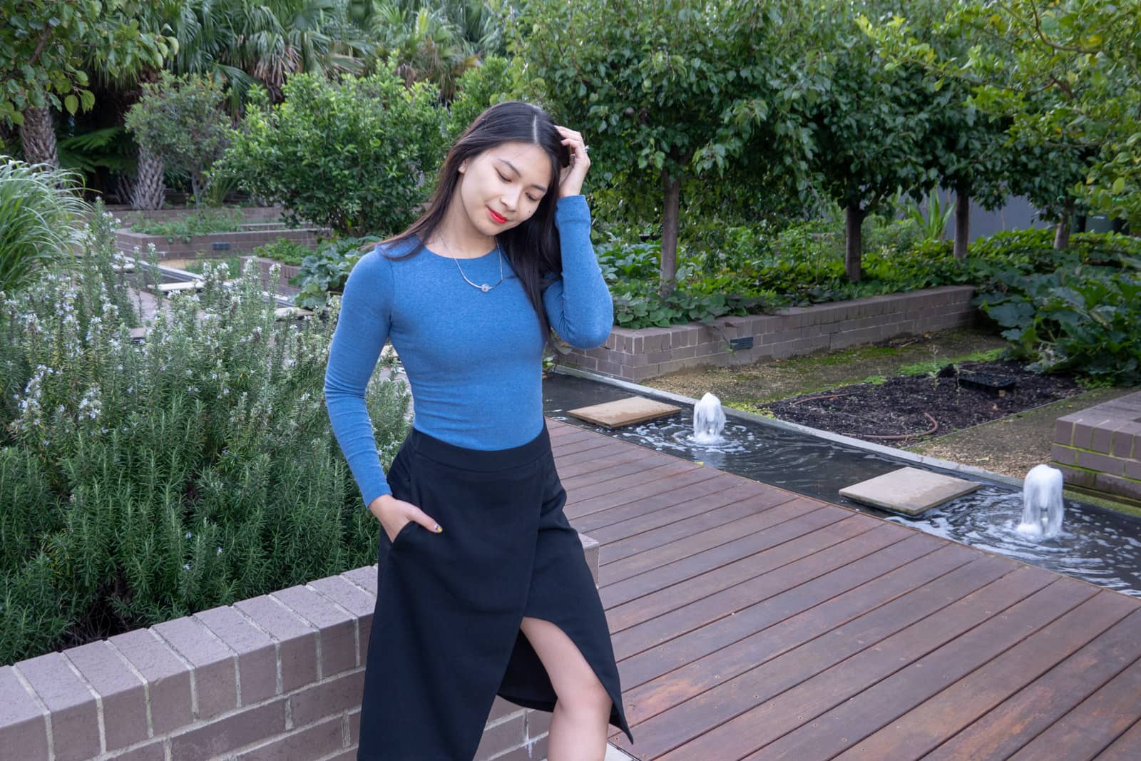 The same woman in other photos on this page, with her eyes downcast and a hand in her pocket, the other hand brushing her hair aside. She is standing by a short brick wall and there is a small stream of small water fountains in the background