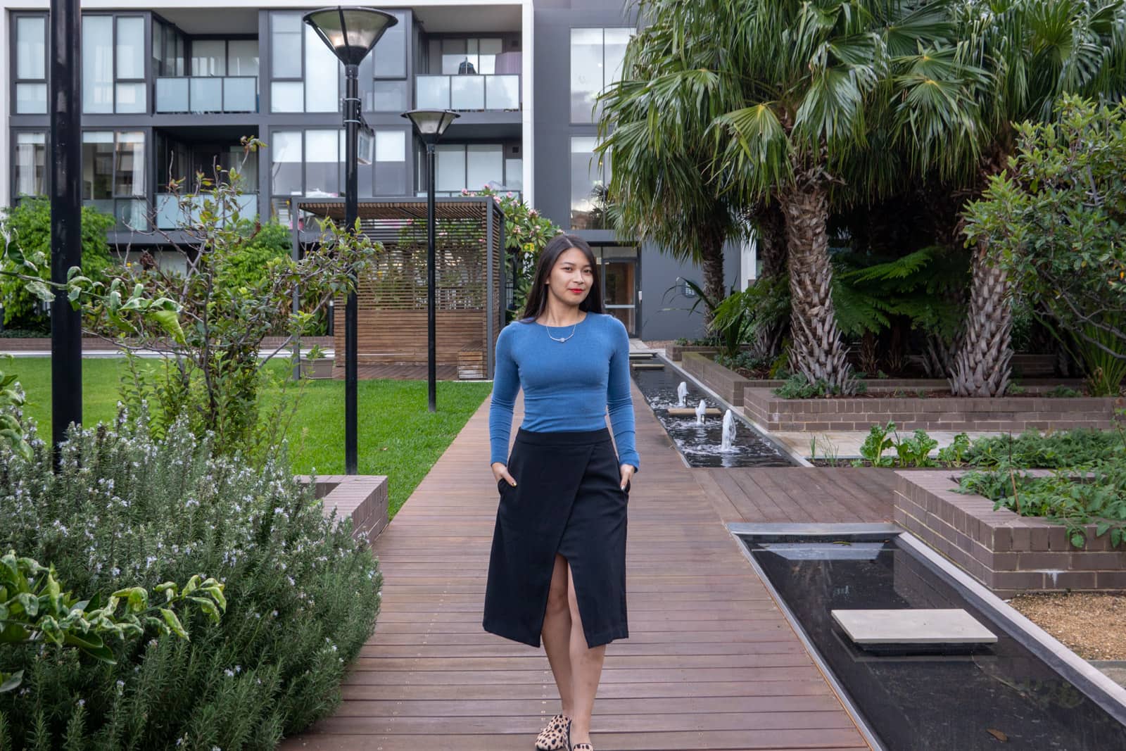 The same woman in other photos on this page, wearing the same outfit. She is standing facing the camera and has her hands in her skirt pockets. She’s standing on a wooden pathway with brick-bordered gardens in the background, and an apartment building with frosted glass balconies