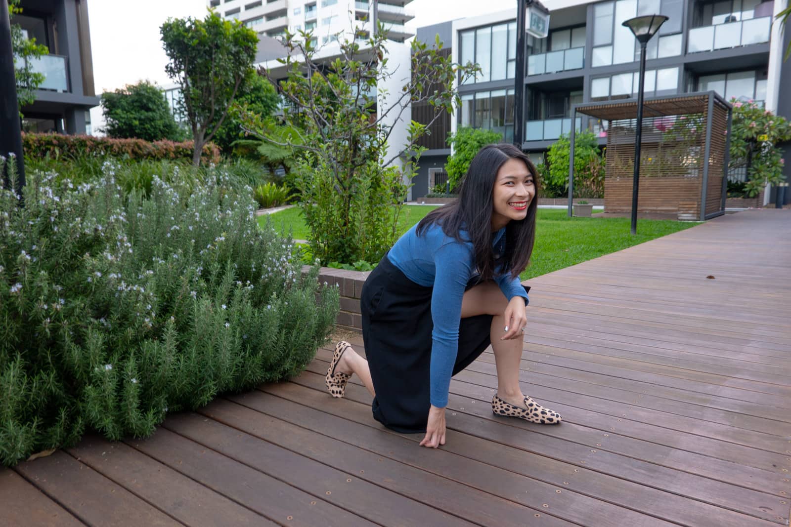 The same woman in other photos in this post, but posing on a wooden plank path with one knee on the ground and the other leg propped up. She is grinning, mid-laugh. In the background a cabana and a block of apartments can be seen