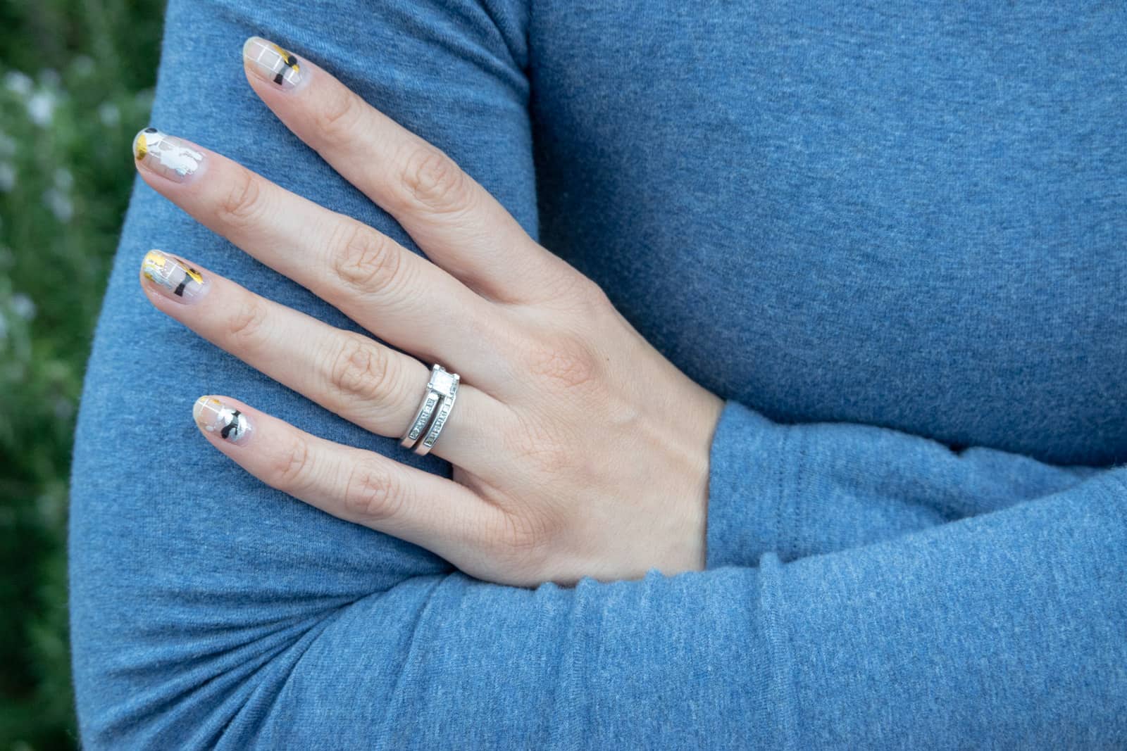 A close up of a woman’s hand touching her opposite upper arm, as her arms are folded. She is wearing a long sleeved blue top. Her nails have silver, gold, black and white nail art on them resembling paint splatters. She has a wedding ring and an engagement ring on her finger.