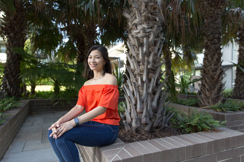 A girl in an orange top and dark blue jeggings, sitting on a small brick wall that forms part of a tree plantation. She is resting he hands on her knees.