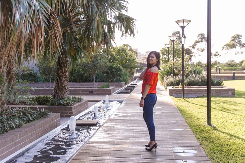 A girl in a park standing with her back slightly to the camera, head turned to the left to face the camera. She is wearing an orange top and dark blue jeggings. The background is a wooden walkway with a small strip of water down the side, with small water bubblers. Lampposts line the other side of the walkway.