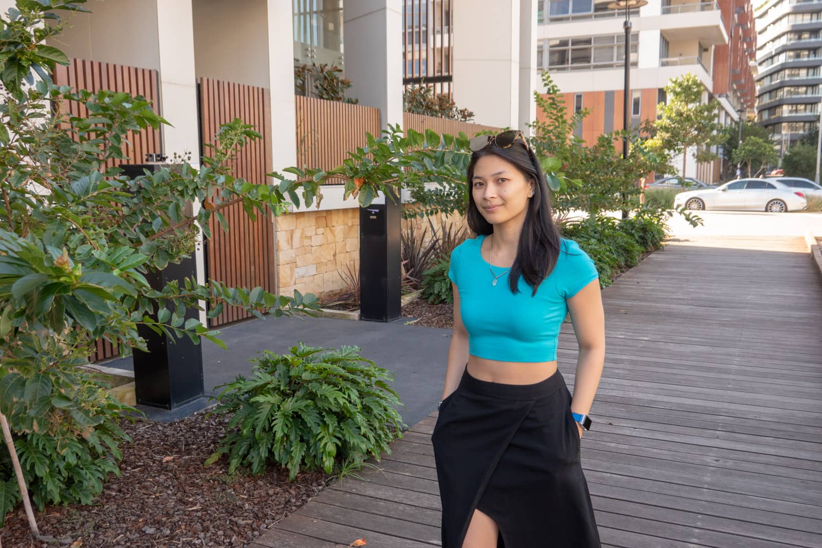 The same woman in other photos on this page, wearing the same outfit. She has her hands in her pockets and is standing on a wooden path. There are apartment buildings in the background