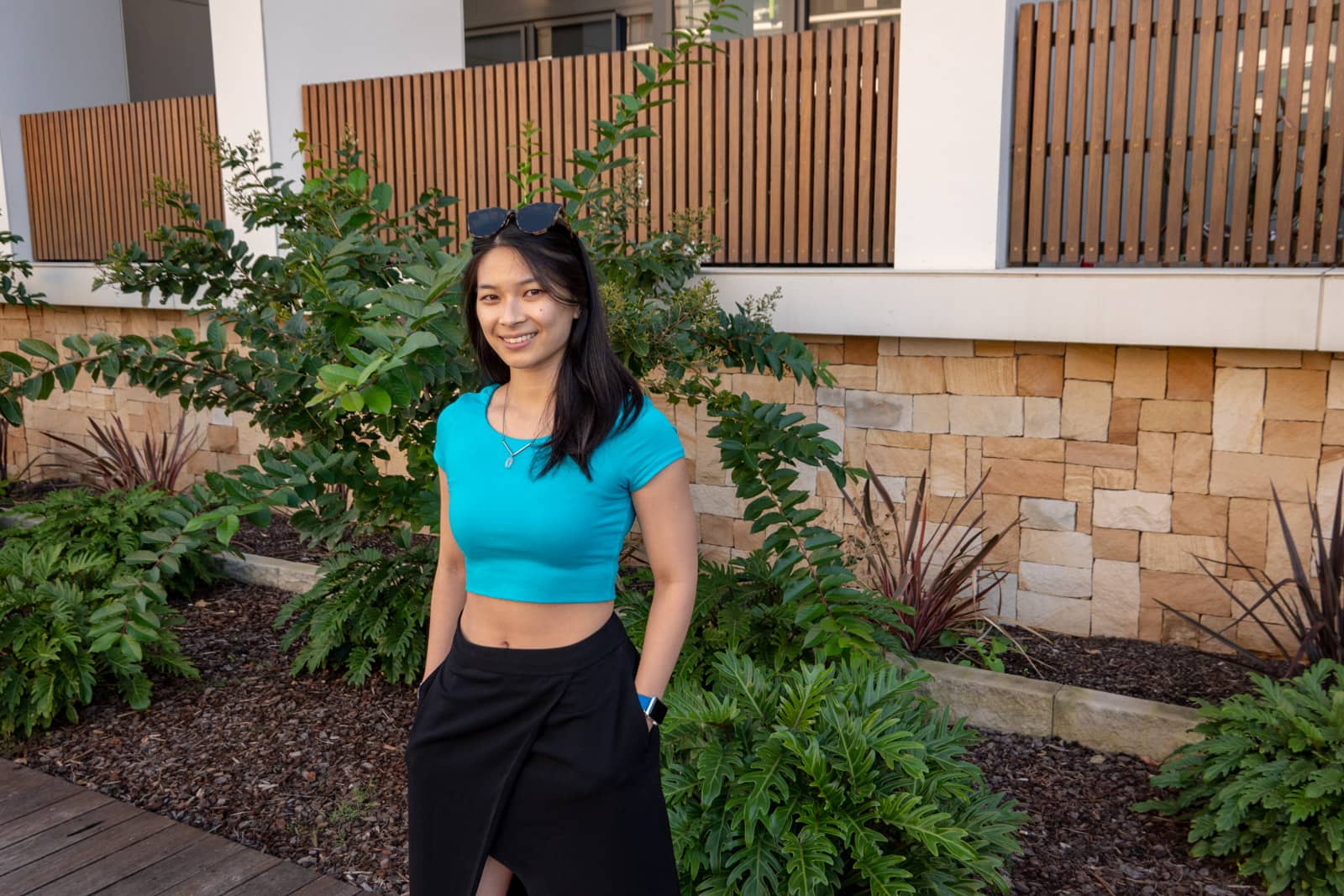 The same woman in other photos on this page, wearing the same outfit. She has her hands in her pockets and is standing by a brown brick wall behind a small patch of garden plants