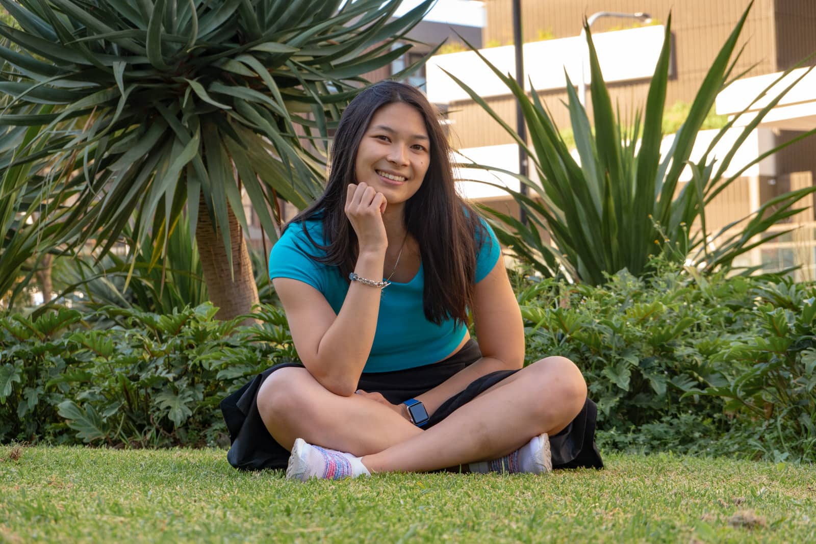 A woman with medium length dark hair, wearing a turquoise tee and a black skirt, sitting cross-legged on the grass. There are some spiky plants behind her. She is resting her chin on her hand and the elbow of that arm is propped on one of her knees.