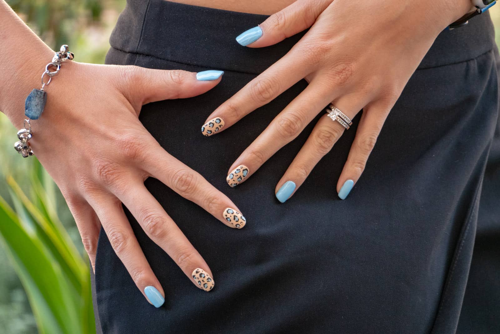 A close-up of a woman’s hands at her hip, with her fingers spread out. Her fingernails are painted blue with some of them having a leopard print pattern. She wears a silver chain with a blue stone on one wrist.