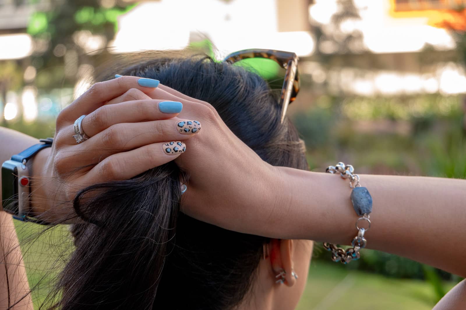 A close-up view of a woman’s head from behind. Her hands are holding her dark hair up in a ponytail. She has blue nails with a few nails having leopard-print nail art on them