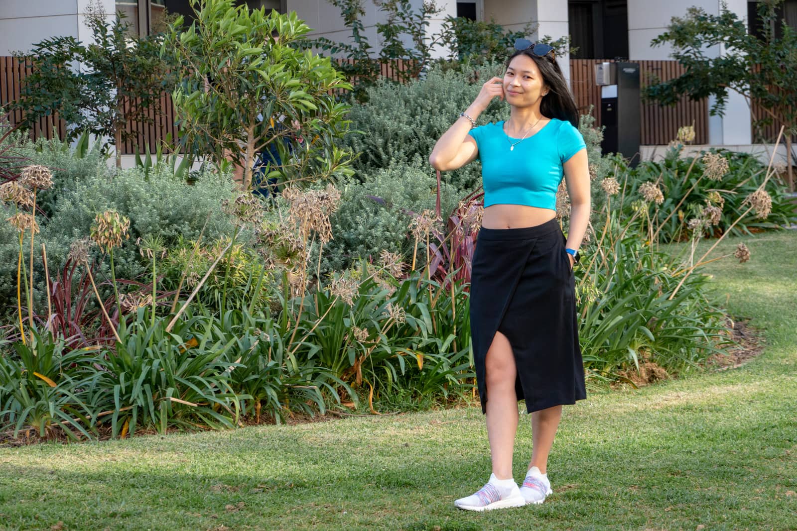 A woman with short dark hair, brushing some hair behind her ear. She is wearing a turquoise top and a long black skirt with white running shoes. She’s standing in a garden