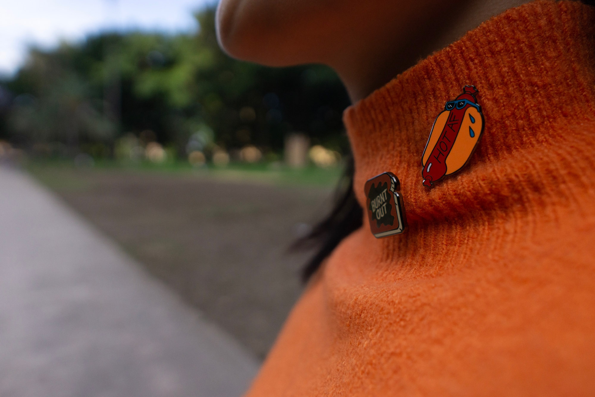 A close up of an orange turtleneck sweater worn by a woman. She is wearing two pins on her sweater; one a hot dog with the text “HOT AF” and the other a piece of toast reading “Burnt out”