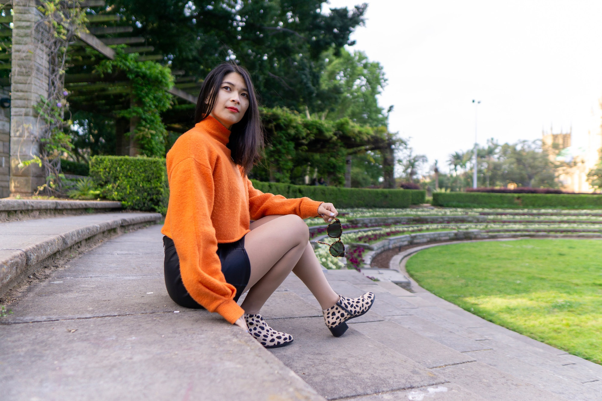 A woman sitting on stone steps. She is wearing a bright orange sweater and a black skirt. Her shoes are light in colour with black spots, resembling giraffe print. She is looking away from the camera and holding a pair of sunglasses in her left hand