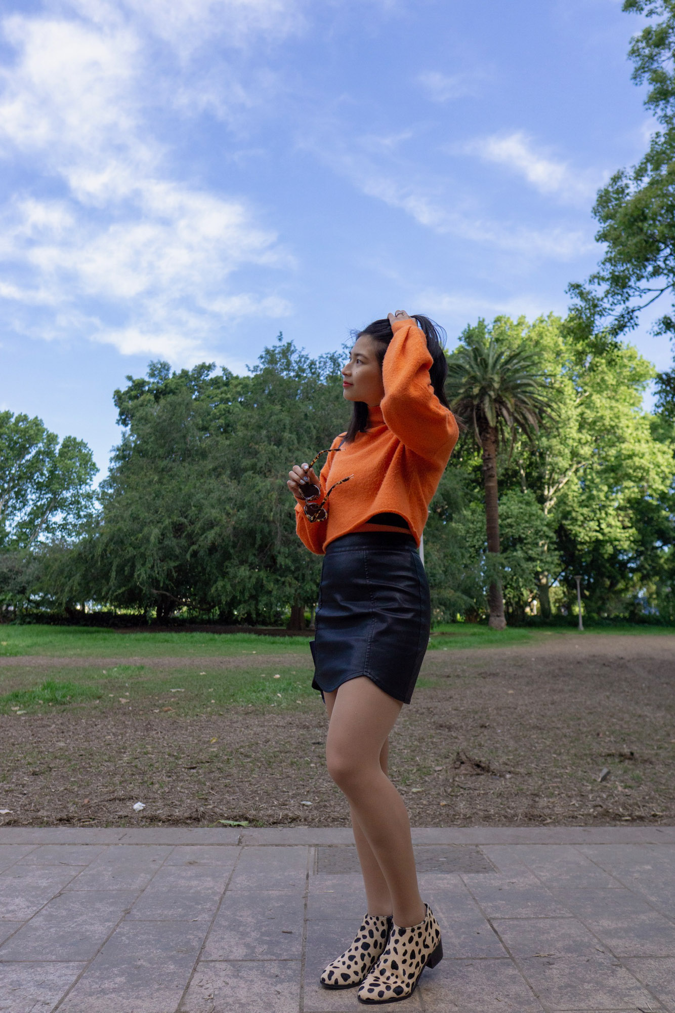 A woman with short dark hair, standing in a park during the day. She is wearing a bright orange sweater and a black skirt. Her boots have black spots resembling a giraffe print