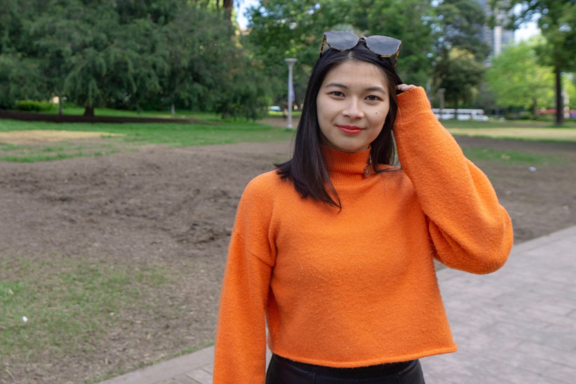 A medium shot of a woman wearing an orange turtleneck sweater. She is tucking hair behind her ears and she has sunglasses on her head