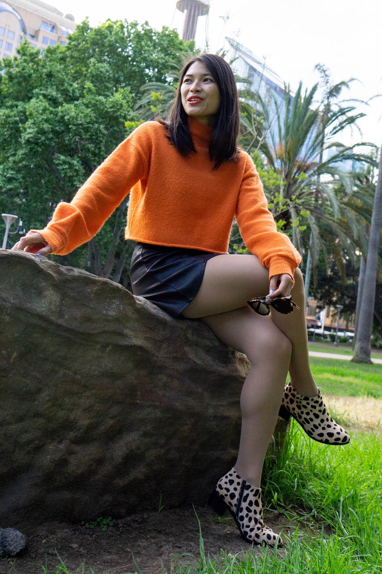 A woman with short dark hair, sitting on a large rock. She is wearing a bright orange sweater and a black skirt. She is in a park and there are skyscrapers behind her.