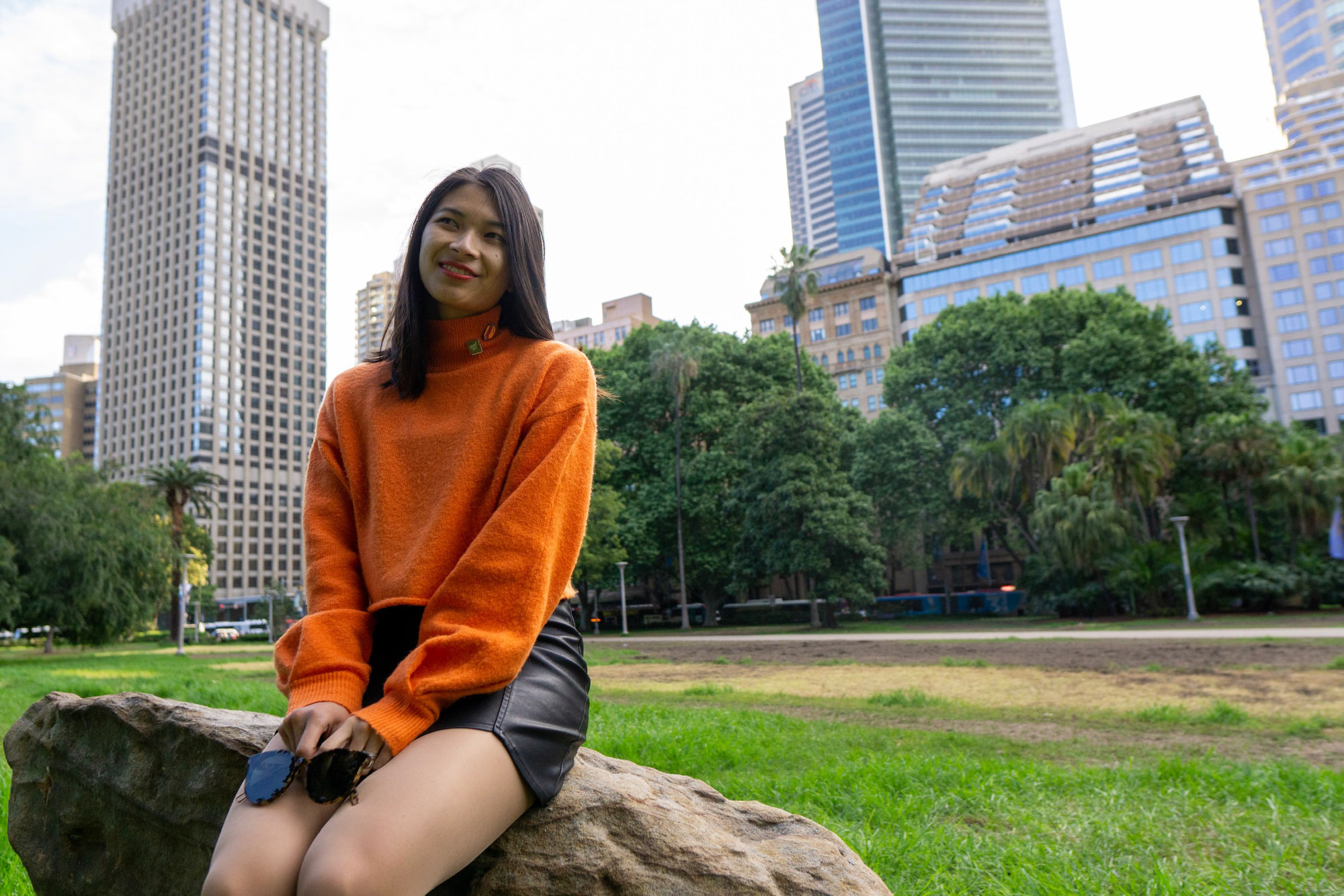 A woman with short dark hair, sitting on a large rock. She is wearing a bright orange sweater and a black skirt. She is in a park and there are skyscrapers behind her.