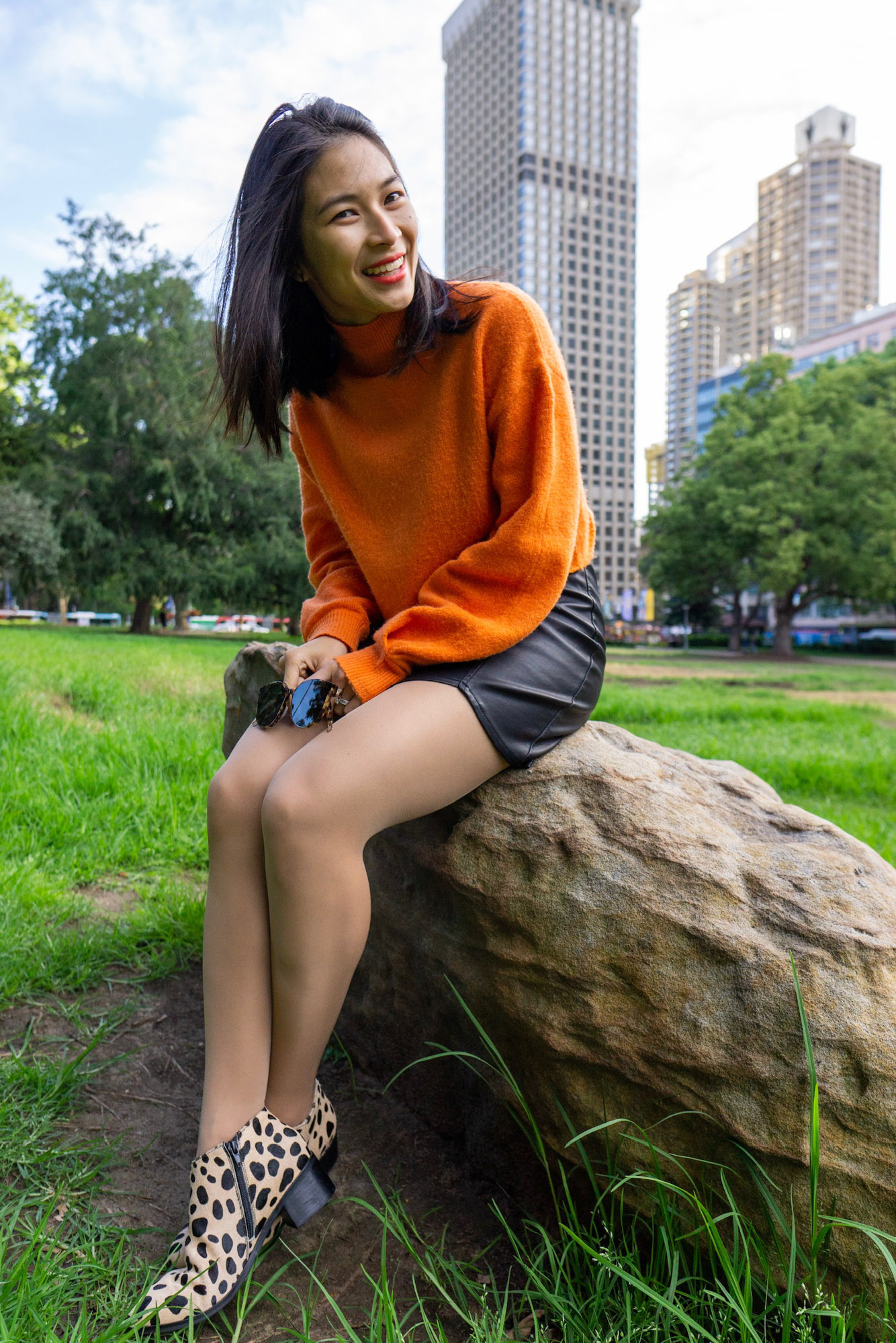 A woman with short dark hair, sitting on a large rock. She is wearing a bright orange sweater and a black skirt. She is in a park and there are skyscrapers behind her.