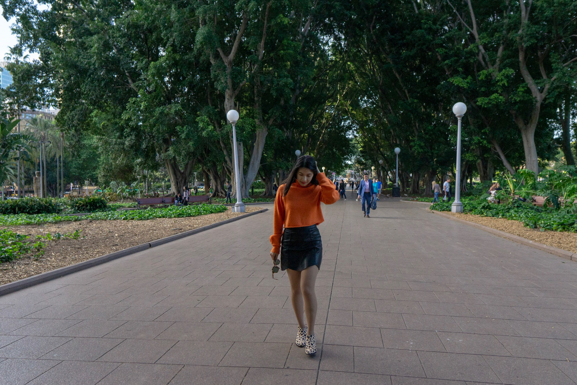 A woman with short dark hair, walking down a pathway in a park with lots of green trees. She is wearing a bright orange sweater and a black skirt.