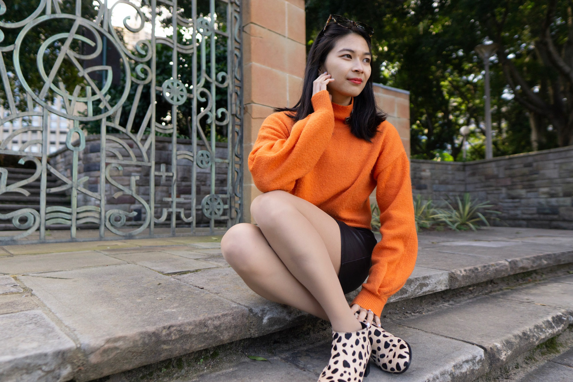 A woman sitting on stone steps. She is wearing a bright orange sweater and a black skirt. Her shoes are light in colour with black spots, resembling giraffe print. Behind her is a metal gate in a pattern.