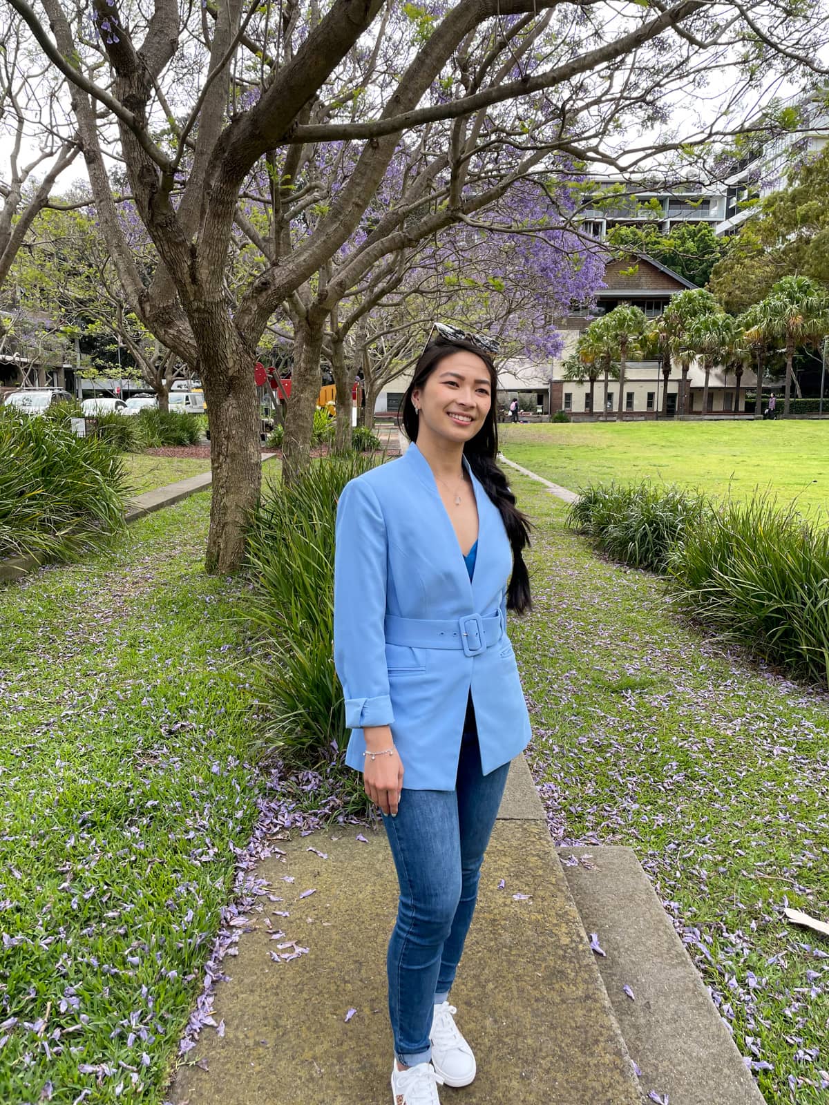 A woman with fair skin and long dark hair braided loosely in a side braid. She is wearing a light blue blazer and blue jeans with white sneakers and is standing in a park.