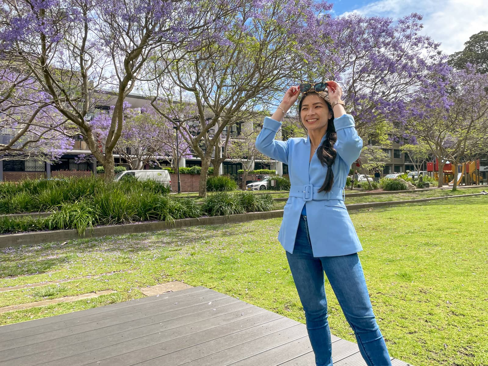 A woman with fair skin and long dark hair braided loosely in a side braid. She is wearing a light blue blazer and blue jeans and is holding her sunglasses on top of her head. She is grinning. In the background are jacaranda trees in a park