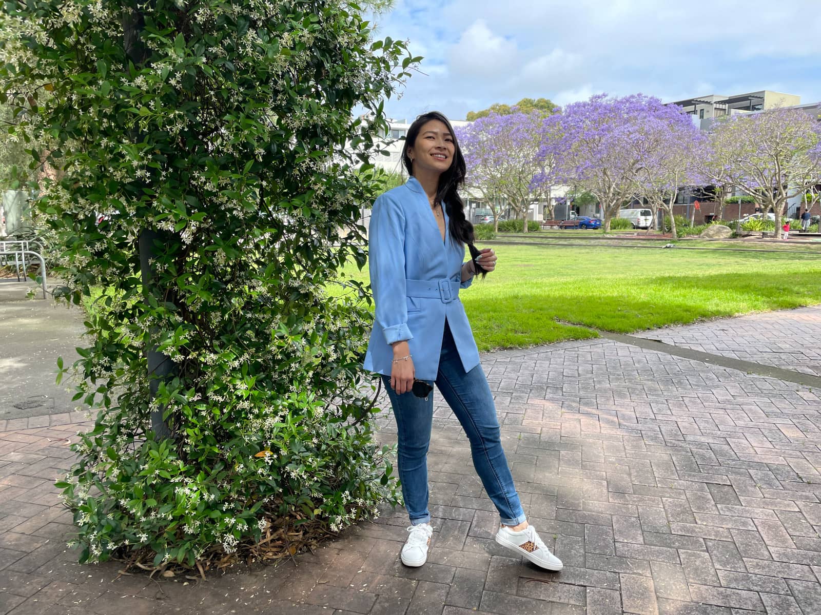 A woman with fair skin and long dark hair braided loosely in a side braid, which she is clutching with one hand. She is wearing a light blue blazer and blue jeans with white sneakers. She is standing at the side of an open park and there is a wire pillar next to her covered in leaves