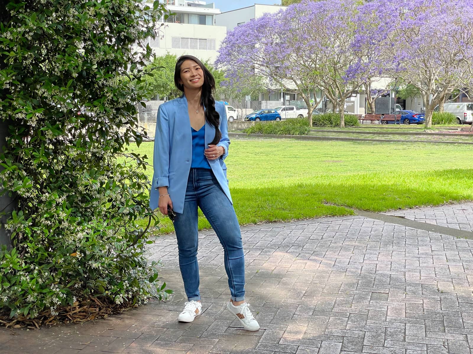 A woman with fair skin and long dark hair braided loosely in a side braid. She is wearing a light blue blazer and blue jeans with white sneakers, and holding onto the open edge of her blazer with one hand, and a pair of sunglasses in the other hand. She is standing at the side of an open park and there is a wire pillar next to her covered in leaves