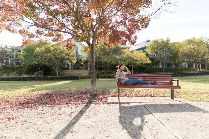 Me sitting sideways on a park bench with a tree shedding autumn-coloured leaves