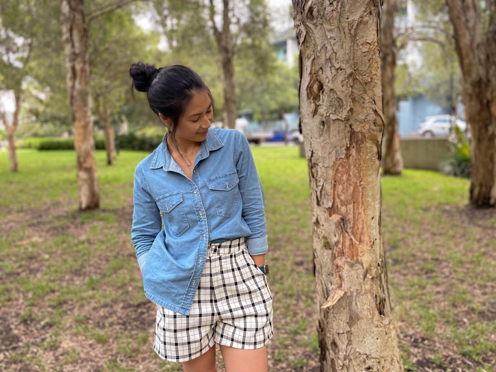 A woman wearing a chambray button-up shirt, half tucked into a pair of shorts with a black and white check pattern. She has her hands in her pockets. Her dark hair is up in a bun and she is looking down and to the side, not facing the camera. She is standing amongst a handful of trees spaced apart.