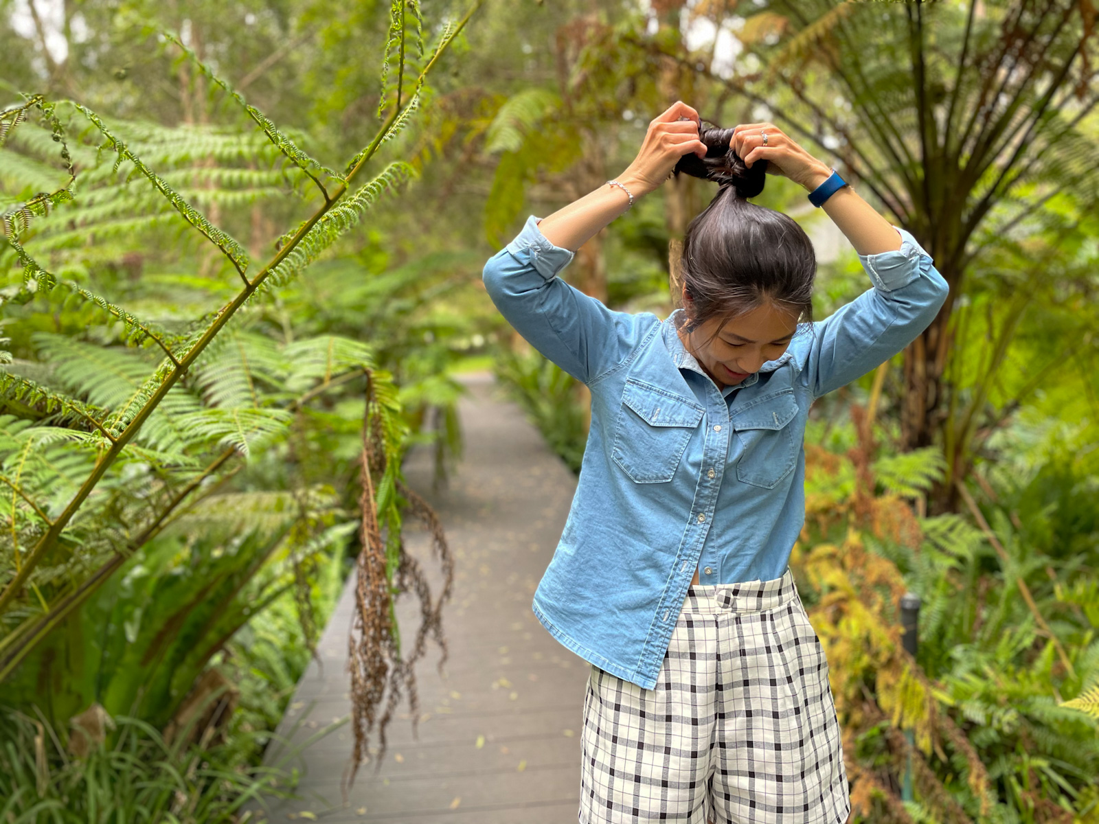 The same woman in previous photos in this post, wearing the same outfit. She is looking down and smiling, and her hands are above her head, adjusting her bun of hair. She is standing on a boardwalk.