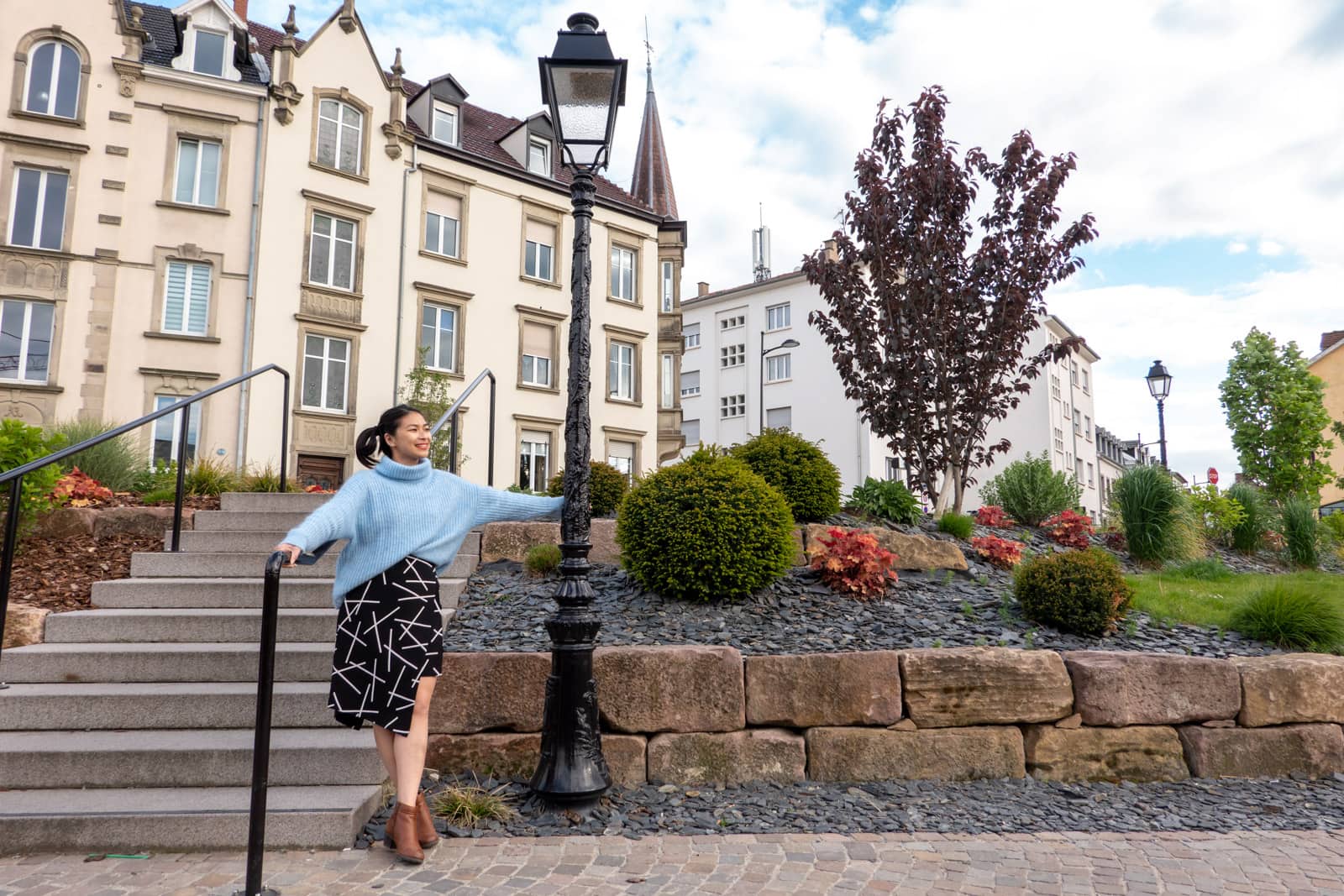 A woman in a blue sweater and a black and white patterned skirt, next to a set of stairs. She’s holding onto a lamppost and in the background is a nice garden and some old buildings with tall windows