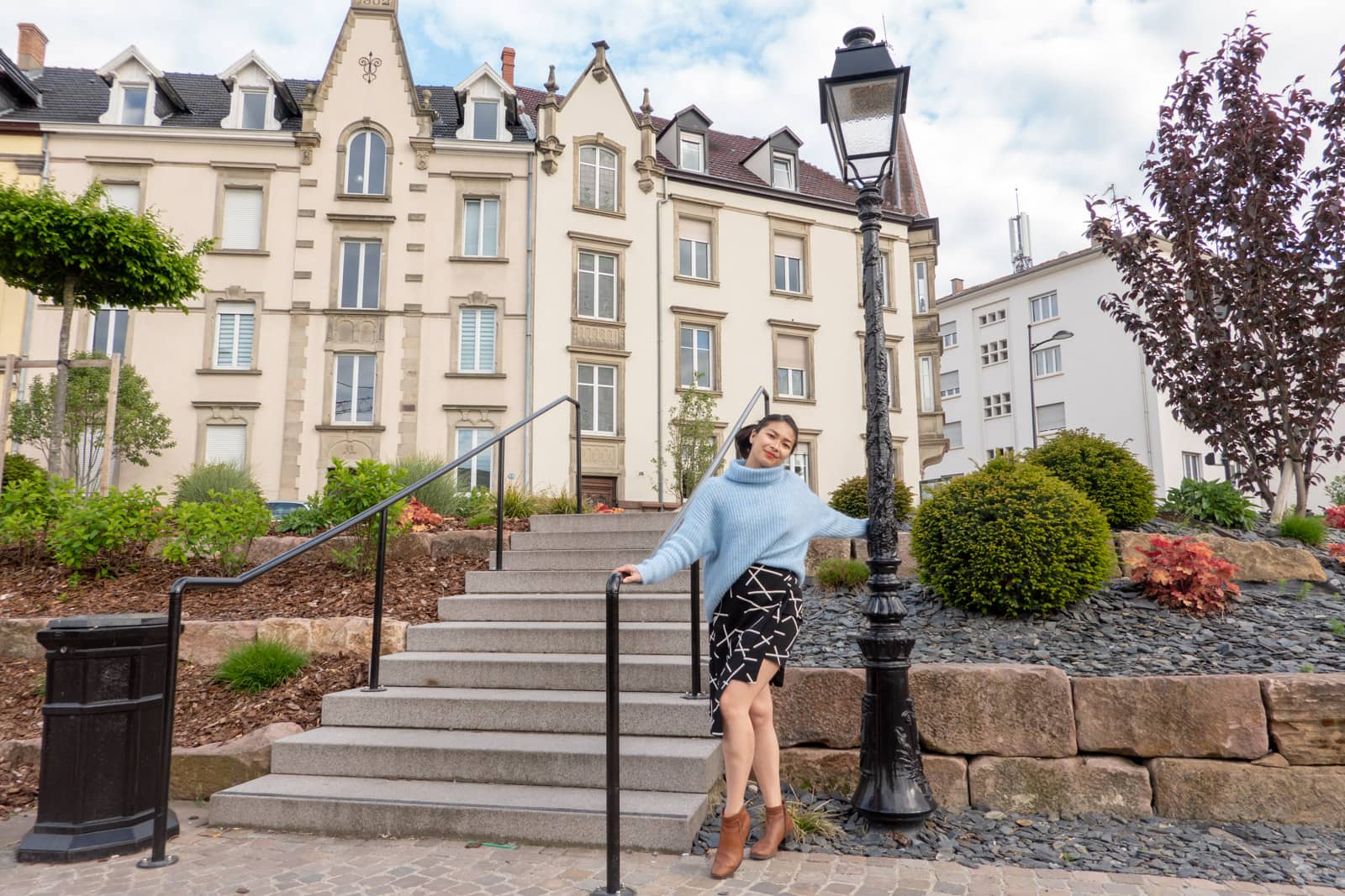 The same woman in other photos on this page, dressed in the same clothes. She’s holding the lamppost but also has her other hand resting on the railing of the stairs.