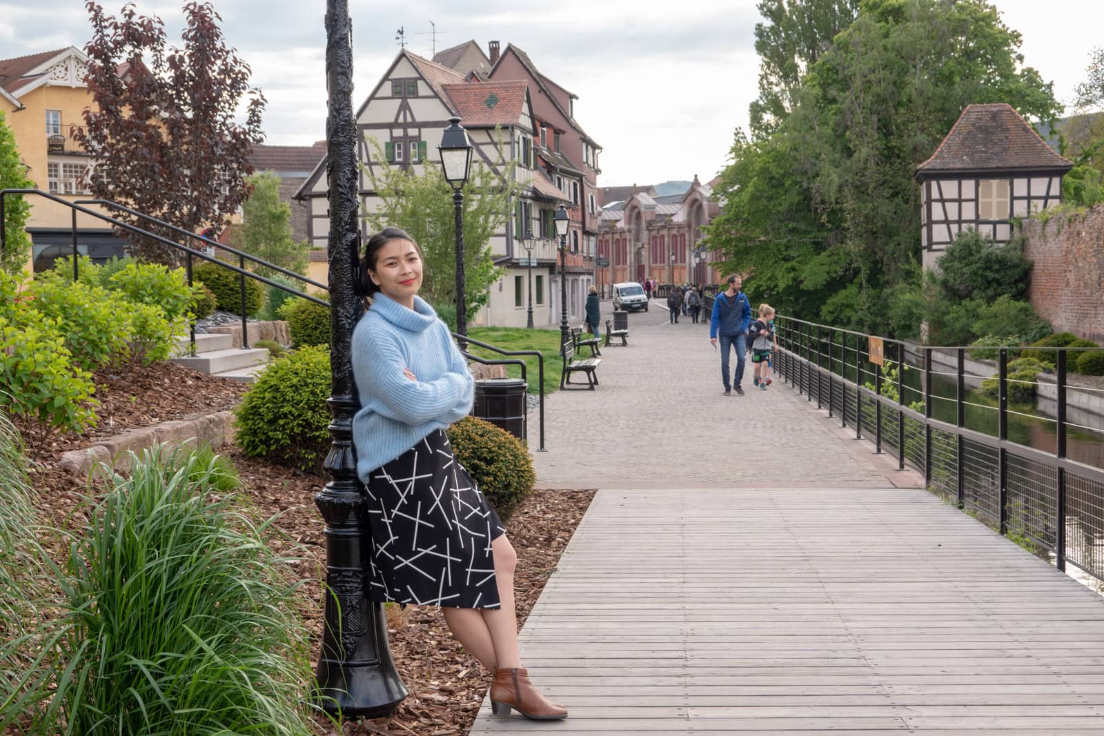 The same woman in other photos on this page, dressed in the same clothes. She is leaning against a lamppost and in the background are the buildings of a French village