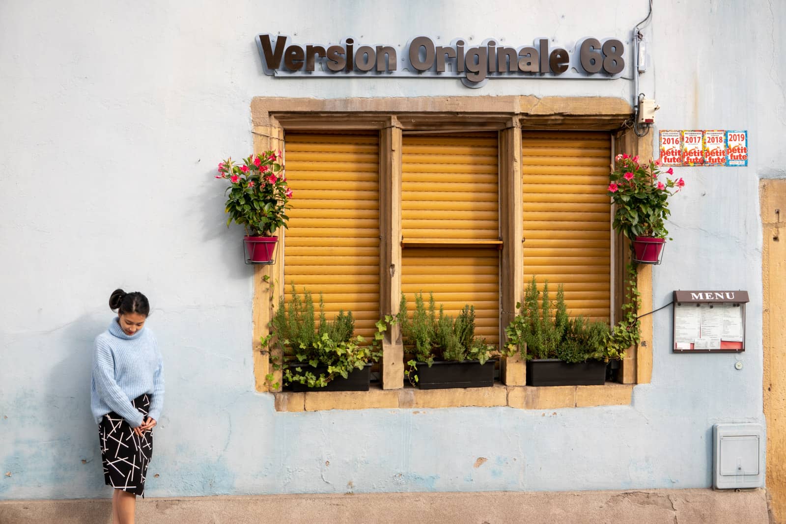 The same woman in other photos on this page, dressed in the same clothes. She is looking down with her hands clasped, standing by a building’s window with mustard shutters, and the building is painted light blue in a similar colour to the woman’s sweater.