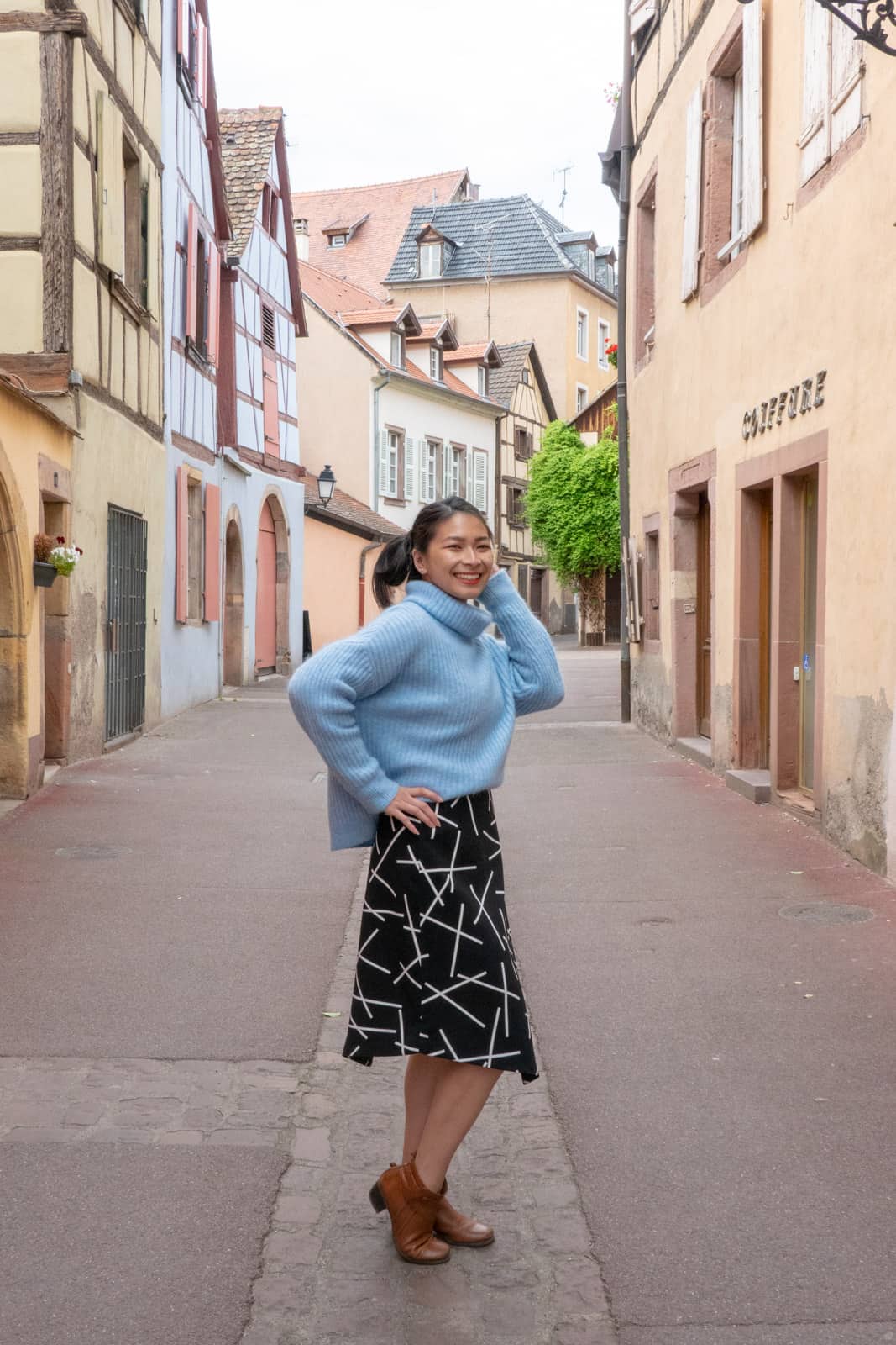 The same woman in other photos on this page, dressed in the same clothes. She has a hand on her hip and the other near her ear, with one knee slightly bent. She is in an alleyway in the afternoon and the buildings around her are peach and pink