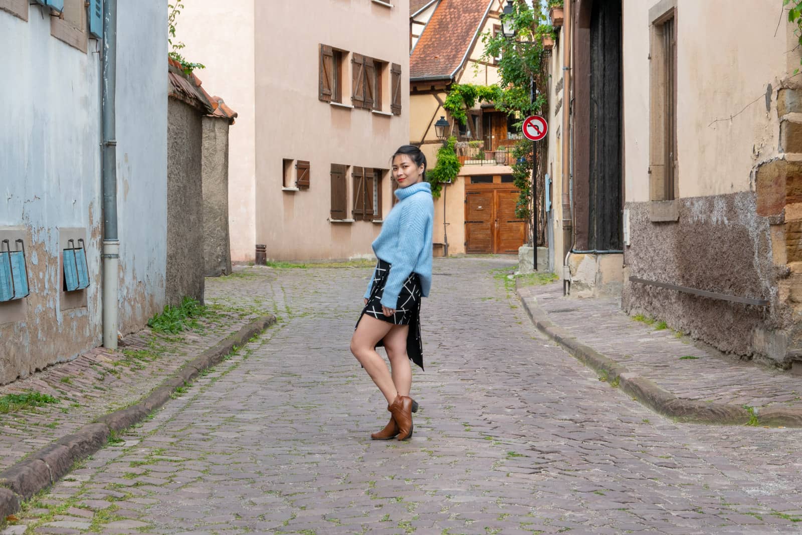 The same woman in other photos on this page, dressed in the same clothes. She is standing side-on, leaning back into her hip with one knee bent. She is in the middle of a quiet cobblestone road.